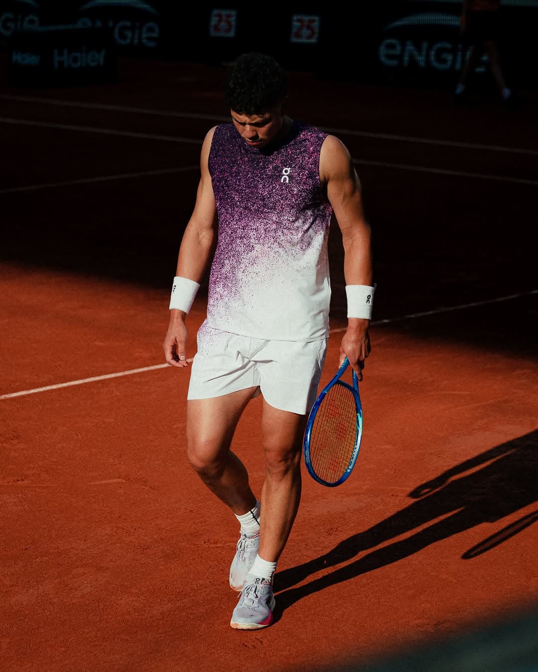Ben walks across red clay court holding blue racquet, wearing purple and white ON kit, match moment captured mid-stride.